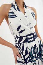 A white midi dress with a floral pattern, featuring a halter neckline, a backless design, and side seam pockets. The dress includes a waist-cinching belt.