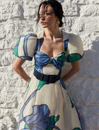A woman wearing a floral printed midi dress with puff sleeves and a sweetheart neckline, standing against a white brick wall.