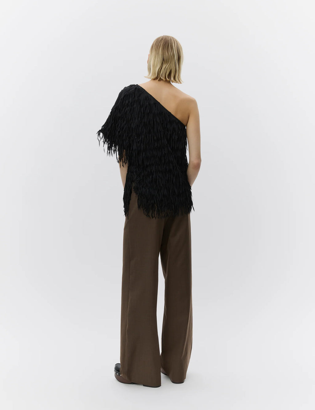 Woman wearing a black one-shoulder feathered top and brown pants on a white background