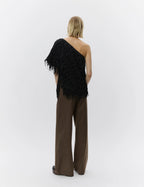 Woman wearing a black one-shoulder feathered top and brown pants on a white background