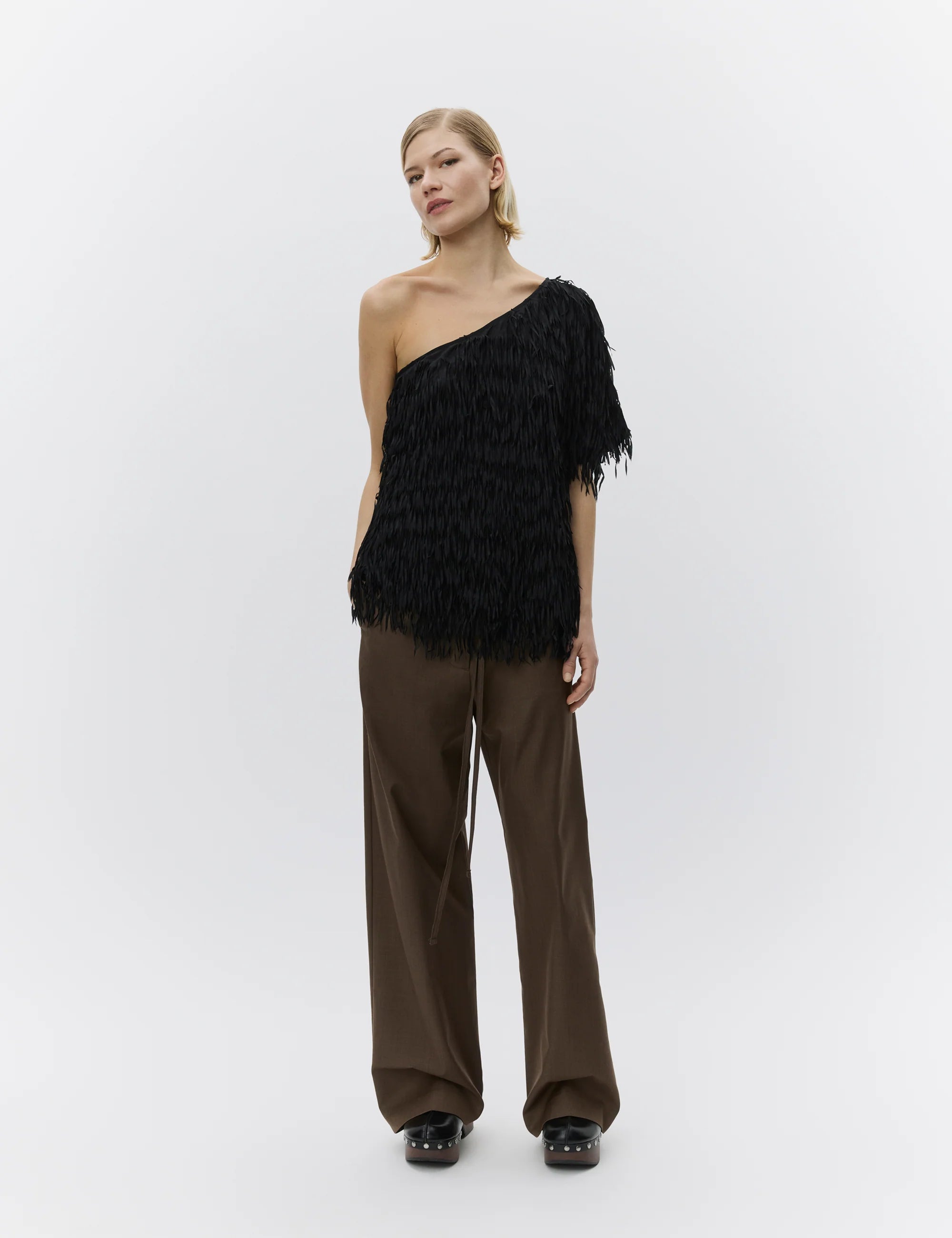 Woman wearing a black one-shoulder feathered top and brown pants on a white background
