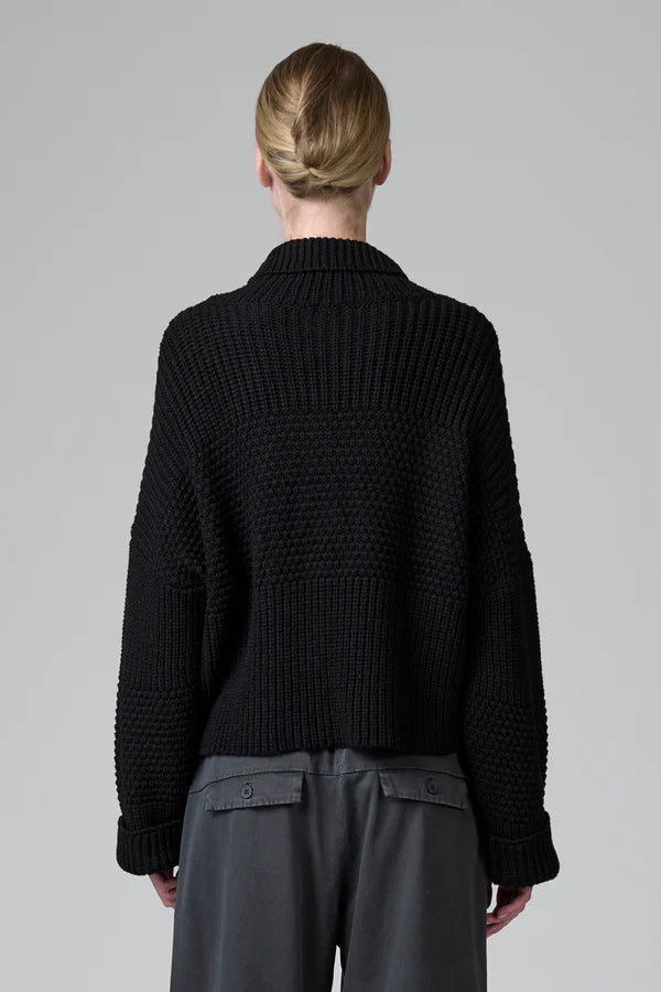 Person wearing a black knitted sweater and gray pants on a plain background