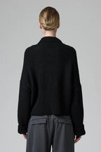 Person wearing a black knitted sweater and gray pants on a plain background