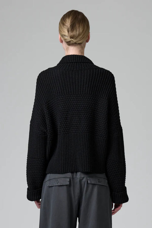 Person wearing a black knitted sweater and gray pants on a plain background