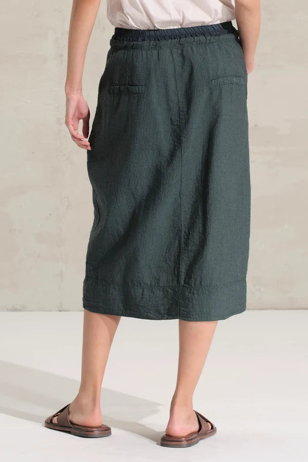 A person shown from the back wearing a dark green linen midi skirt with an elastic waistband and side pockets, paired with a light top and brown sandals, against a neutral concrete wall.