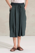 A person wearing dark green wide-leg linen midi skirt with a drawstring waist, paired with brown slide sandals and a white top, standing against a neutral concrete wall.