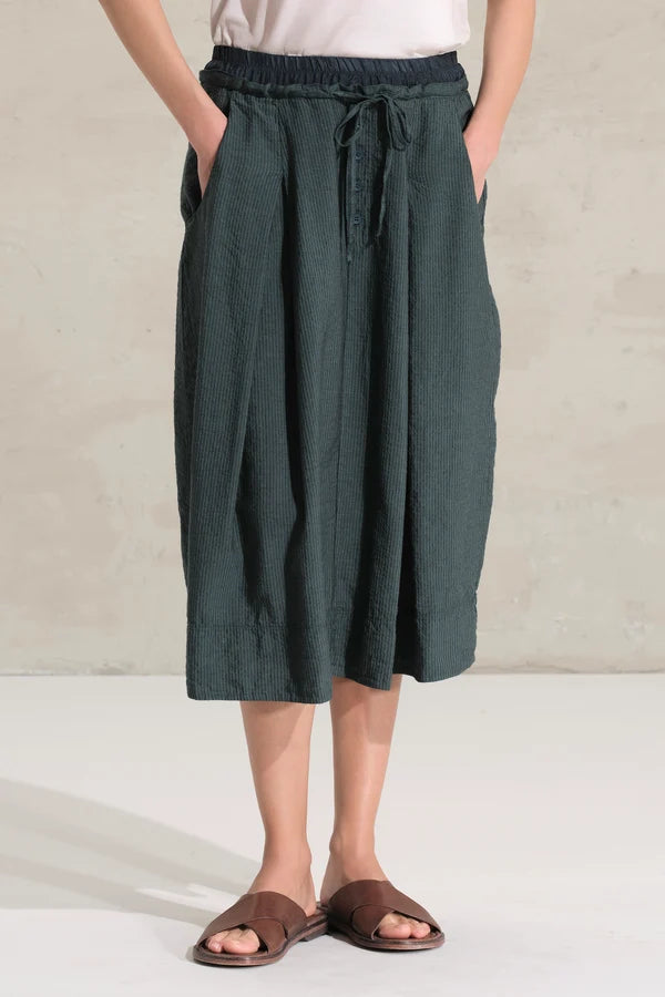 A person wearing dark green wide-leg linen midi skirt with a drawstring waist, paired with brown slide sandals and a white top, standing against a neutral concrete wall.