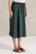 A person wearing a dark green wide-leg linen midi skirt with a drawstring waist, paired with brown slide sandals, standing against a neutral textured beige wall.