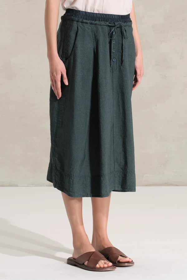 A person wearing a dark green wide-leg linen midi skirt with a drawstring waist, paired with brown slide sandals, standing against a neutral textured beige wall.