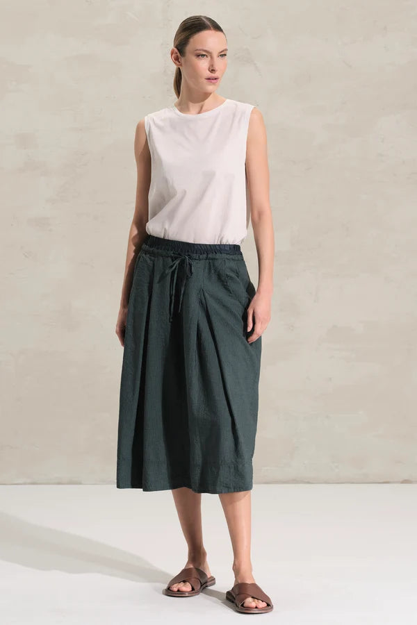 A woman modeling a sleeveless white top and a dark green pleated midi skirt with a drawstring waist, standing against a textured light beige wall.