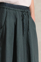 Close-up of a person wearing dark green textured wide-leg pants with an elastic waistband, drawstring, and side buttons, paired with a light sleeveless top.