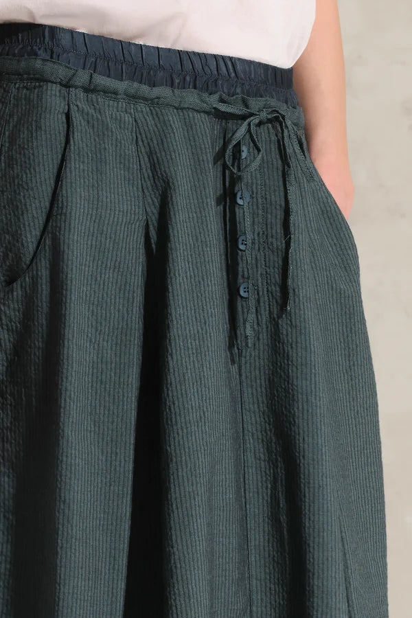 Close-up of a person wearing dark green textured wide-leg pants with an elastic waistband, drawstring, and side buttons, paired with a light sleeveless top.