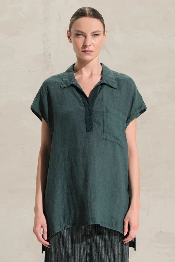 A woman modeling a dark teal-green short-sleeve collared shirt with a partial button placket and a chest pocket, paired with dark striped trousers, against a textured light beige wall.