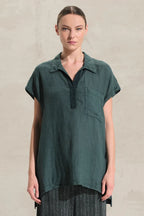 A woman modeling a dark teal-green short-sleeve collared shirt with a partial button placket and a chest pocket, paired with dark striped trousers, against a textured light beige wall.
