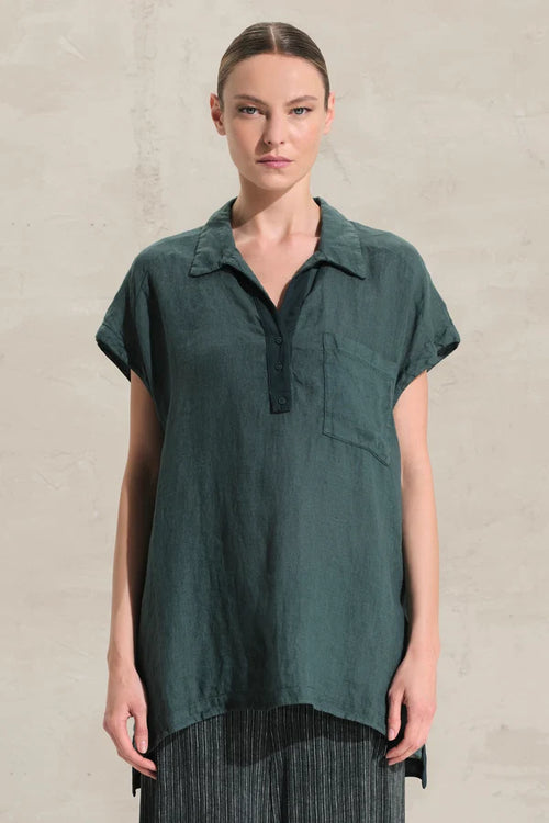 A woman modeling a dark teal-green short-sleeve collared shirt with a partial button placket and a chest pocket, paired with dark striped trousers, against a textured light beige wall.