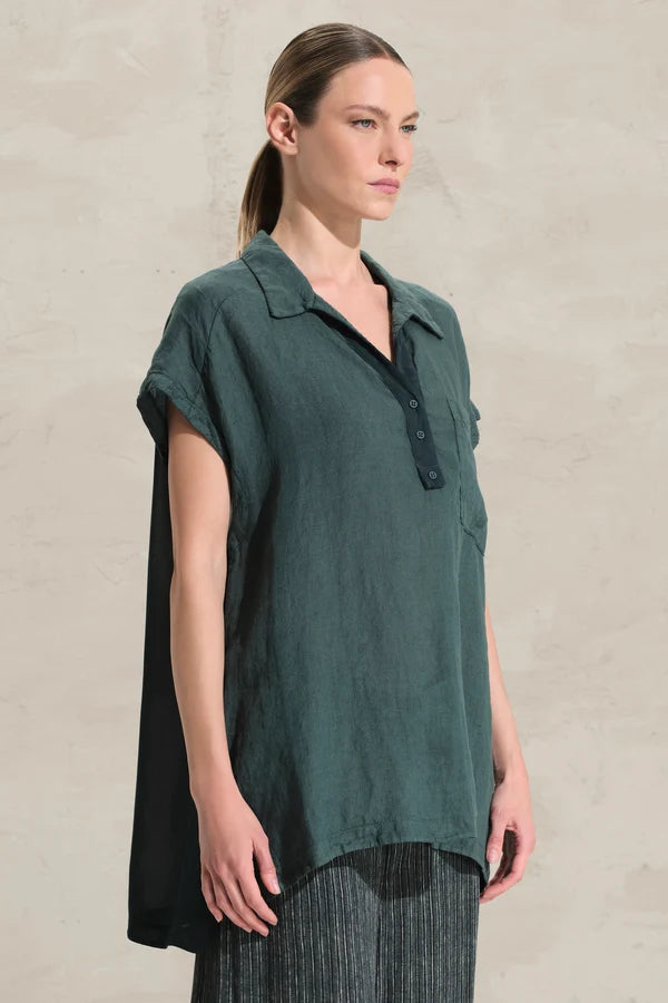 A woman modeling a dark green short-sleeve button-up shirt with a relaxed, oversized fit and a draped side detail, paired with dark gray striped trousers, set against a textured beige wall.