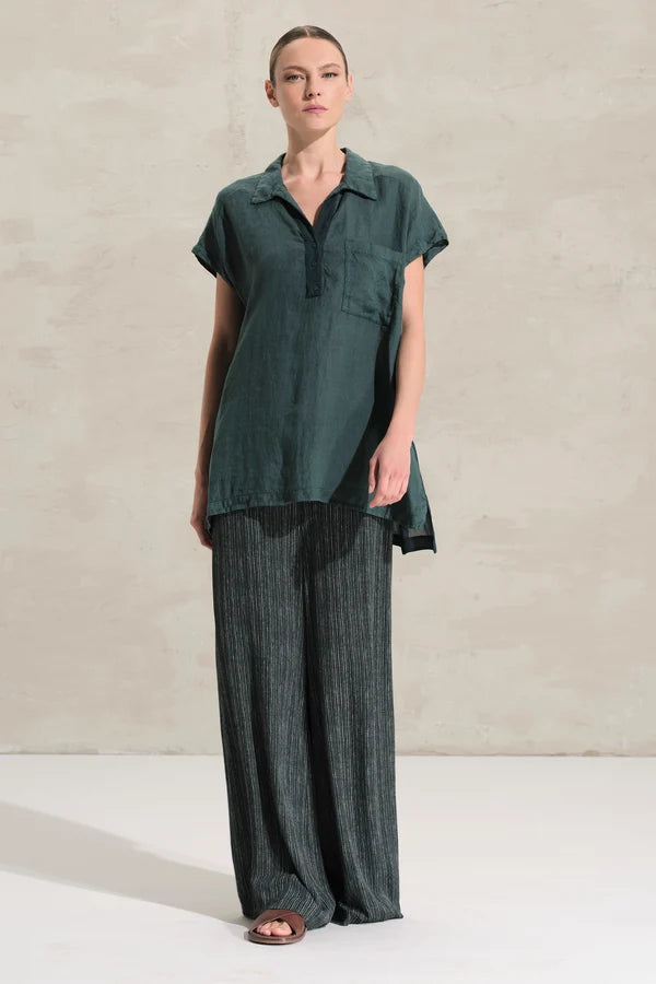 A woman modeling a dark green short-sleeve collared top with a button placket and chest pocket, paired with dark wide-leg pleated trousers, standing against a textured light beige wall.