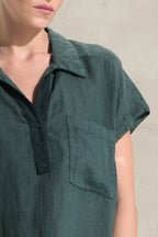 A close-up of a person wearing a dark green short-sleeve collared shirt with a button placket and a chest pocket, against a light neutral wall.