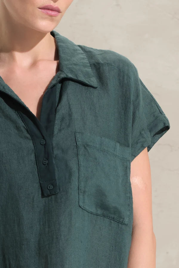 A close-up of a person wearing a dark green short-sleeve collared shirt with a button placket and a chest pocket, against a light neutral wall.