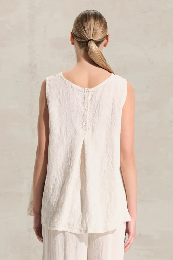 A woman viewed from behind wearing a sleeveless cream-colored linen top with a buttoned back neckline and matching light pants, set against a textured light beige wall.