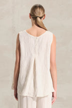 A woman viewed from behind wearing a sleeveless cream-colored linen top with a buttoned back neckline and matching light pants, set against a textured light beige wall.