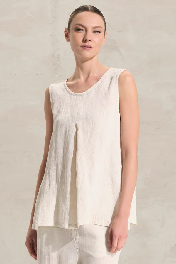 A woman wearing a sleeveless, high-low hem top in a light cream or off-white linen-like fabric, set against a neutral textured wall.