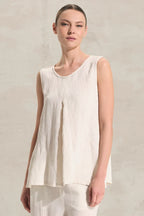 A woman wearing a sleeveless, high-low hem top in a light cream or off-white linen-like fabric, set against a neutral textured wall.