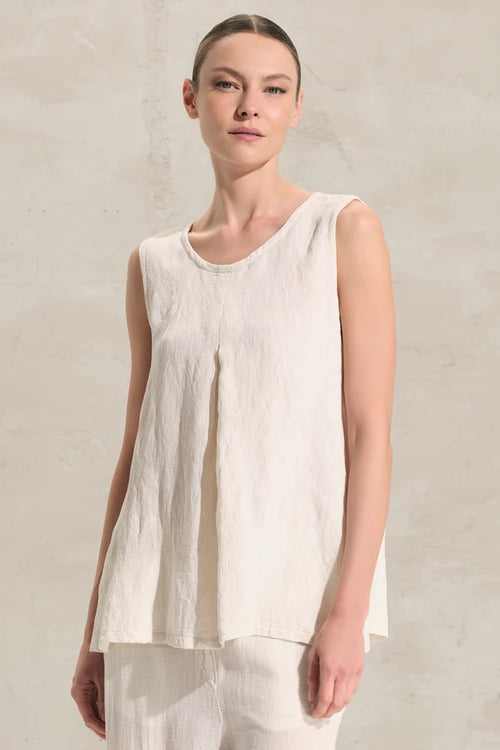 A woman wearing a sleeveless, high-low hem top in a light cream or off-white linen-like fabric, set against a neutral textured wall.