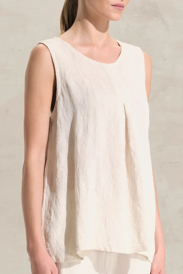 A woman wearing a sleeveless, high-low hem top in a light cream or off-white linen-like fabric, set against a neutral textured wall.