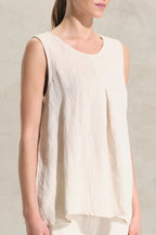 A woman wearing a sleeveless, high-low hem top in a light cream or off-white linen-like fabric, set against a neutral textured wall.