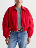 A model wearing a bright red cropped bomber jacket over a gray ribbed crop top and blue jeans, shown from the waist up against a plain white studio background.