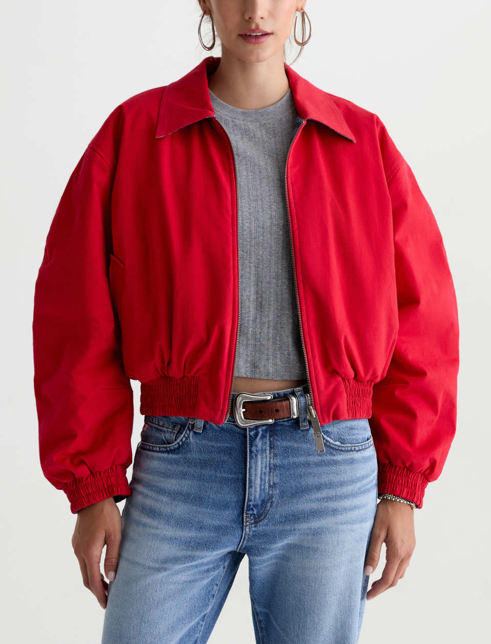 A model wearing a bright red cropped bomber jacket over a gray ribbed crop top and blue jeans, shown from the waist up against a plain white studio background.