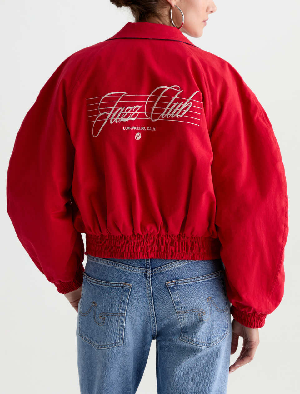 A person shown from the back wearing a bright red satin bomber jacket with 'Jazz Club' script and 'Los Angeles, Calif.' printed on the back, paired with blue jeans against a light gray studio background.