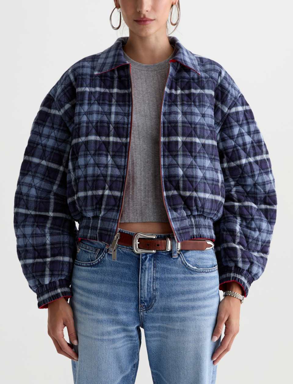 A model wearing a navy and blue plaid quilted jacket with red trim, styled with a gray ribbed top, brown leather belt, and light blue jeans against a plain white background.