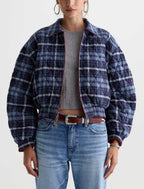 A model wearing a navy and blue plaid quilted jacket with red trim, styled with a gray ribbed top, brown leather belt, and light blue jeans against a plain white background.