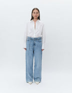 A person wearing a Day Birger et Mikkelsen white button-up shirt with a rounded and dipped hem, and buttoned cuffs, paired with blue jeans.