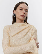 A woman standing with her arms crossed, wearing a beige maxi dress with sequins and a high neck.