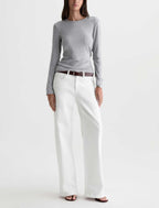A model wearing a light gray long-sleeve knit top, white wide-leg trousers, and a brown leather belt, standing against a plain white studio background.