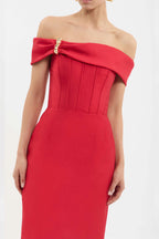 A woman wearing a bright red off-the-shoulder midi dress with a structured corset-style bodice and a decorative gold embellishment on the bow detail at the neckline, photographed against a plain light background.