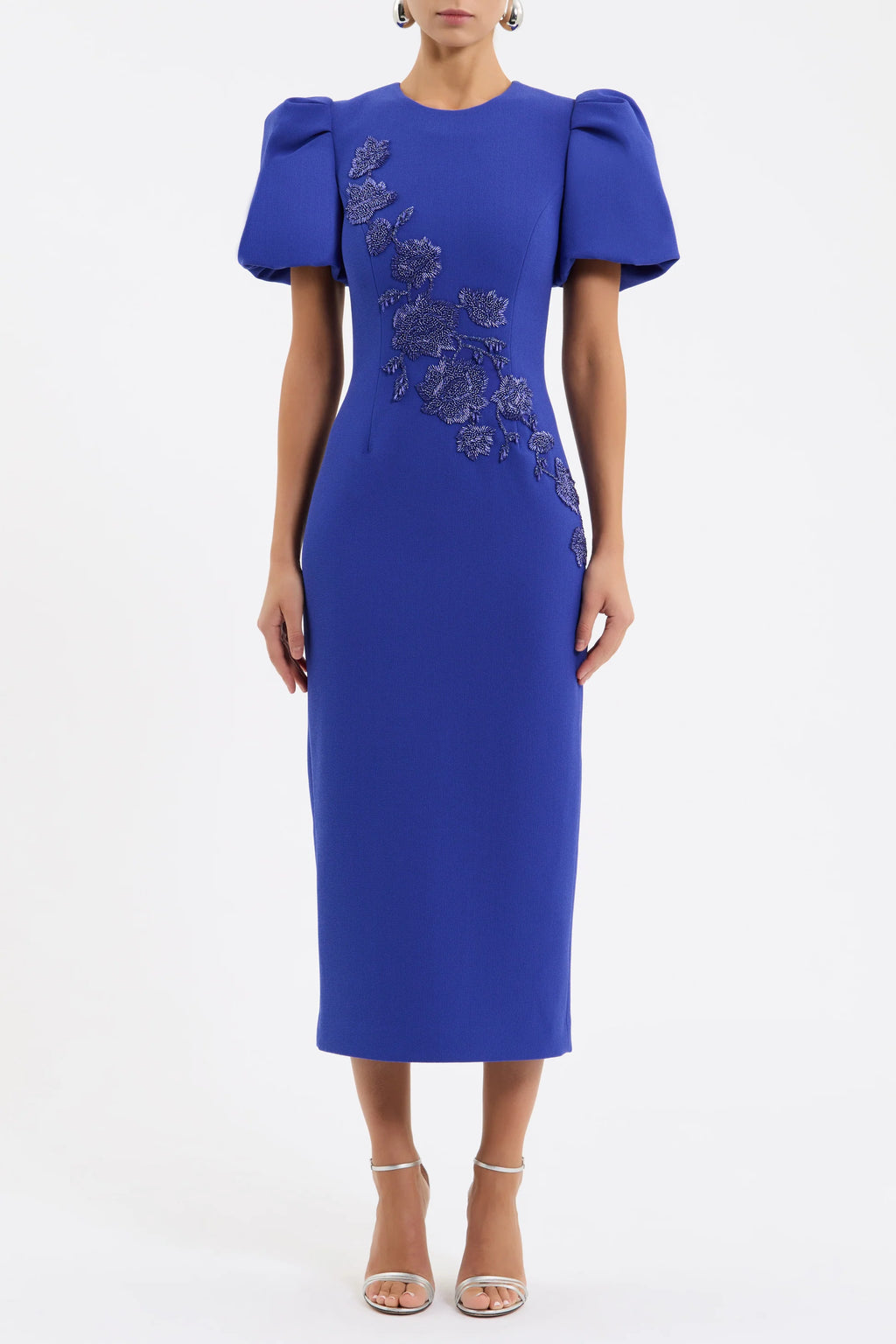 The Rebecca Vallance Evolet Puff Sleeve Midi Dress is captivating in a vibrant ultra marine blue, adorned with intricate floral embellishments. Tailored from bonded crepe, its crew neckline and voluminous short puff sleeves create a refined and elegant shape, complemented by a fitted bodice that flatters the figure. The pencil midi skirt creates an elegant silhouette, detailed with a back split for ease of movement. Secured with an invisible zip fastening at the back and lining for comfort, this dress is a 
