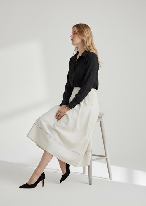 Woman sitting on a stool wearing a black shirt and white skirt against a white background