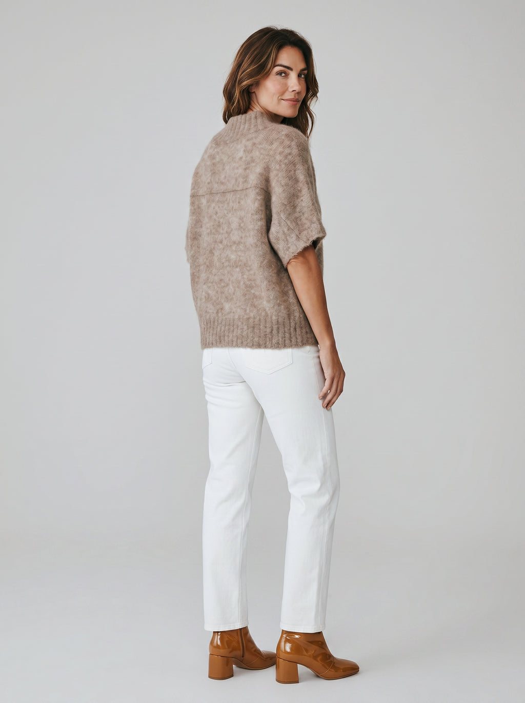 Woman wearing a brown sweater, white pants, and brown boots on a plain background