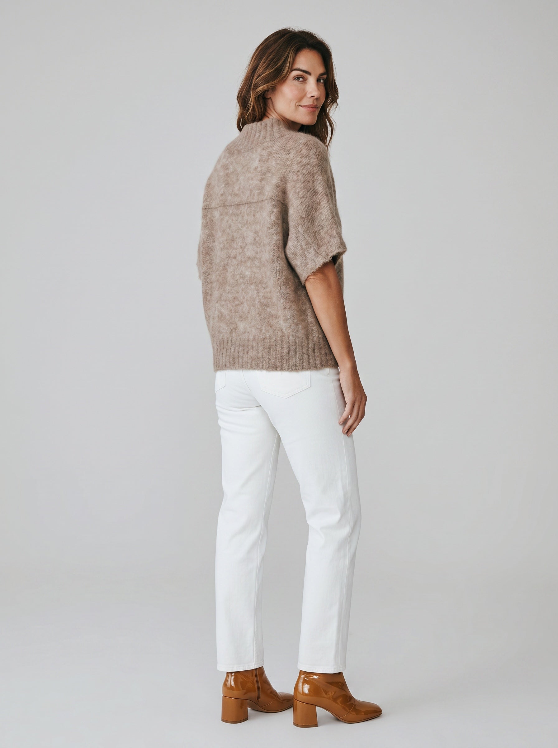 Woman wearing a brown sweater, white pants, and brown boots on a plain background