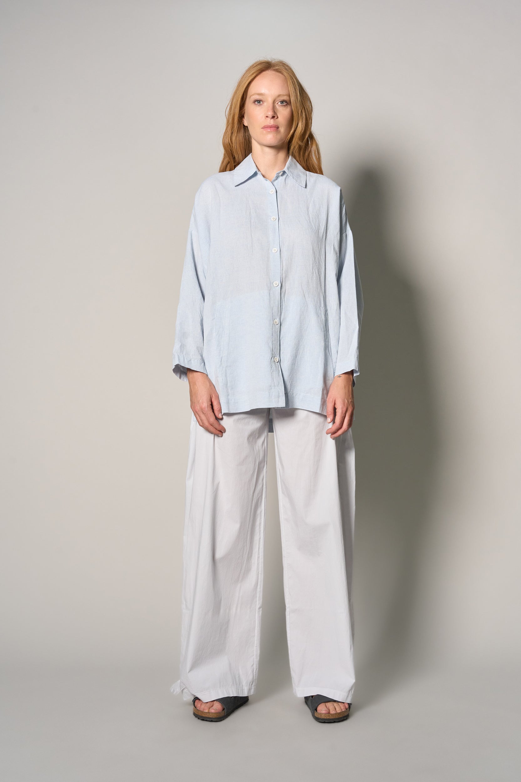 A woman stands against a plain light gray studio background wearing a loose light blue button-up shirt with white wide-leg pants and dark sandals.