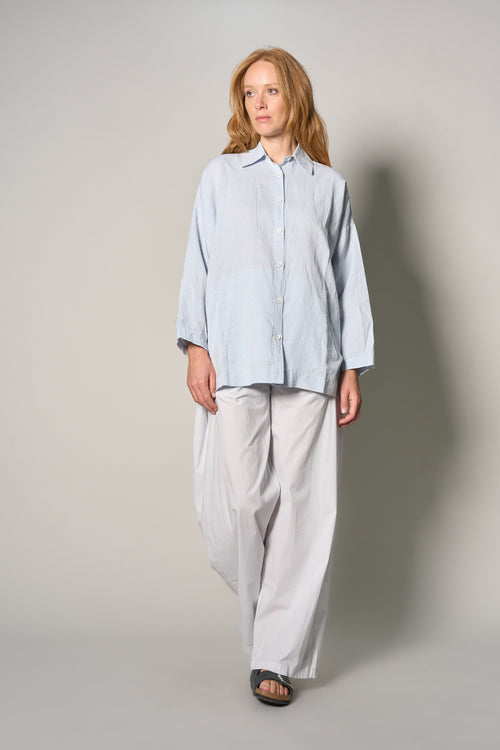 A woman modeling a light blue long-sleeve button-up shirt and loose white wide-leg pants against a neutral gray studio background.