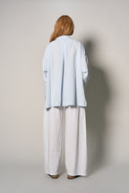 A model viewed from behind wearing a light blue oversized button-down shirt and white wide-leg pants, standing against a neutral light gray studio background.