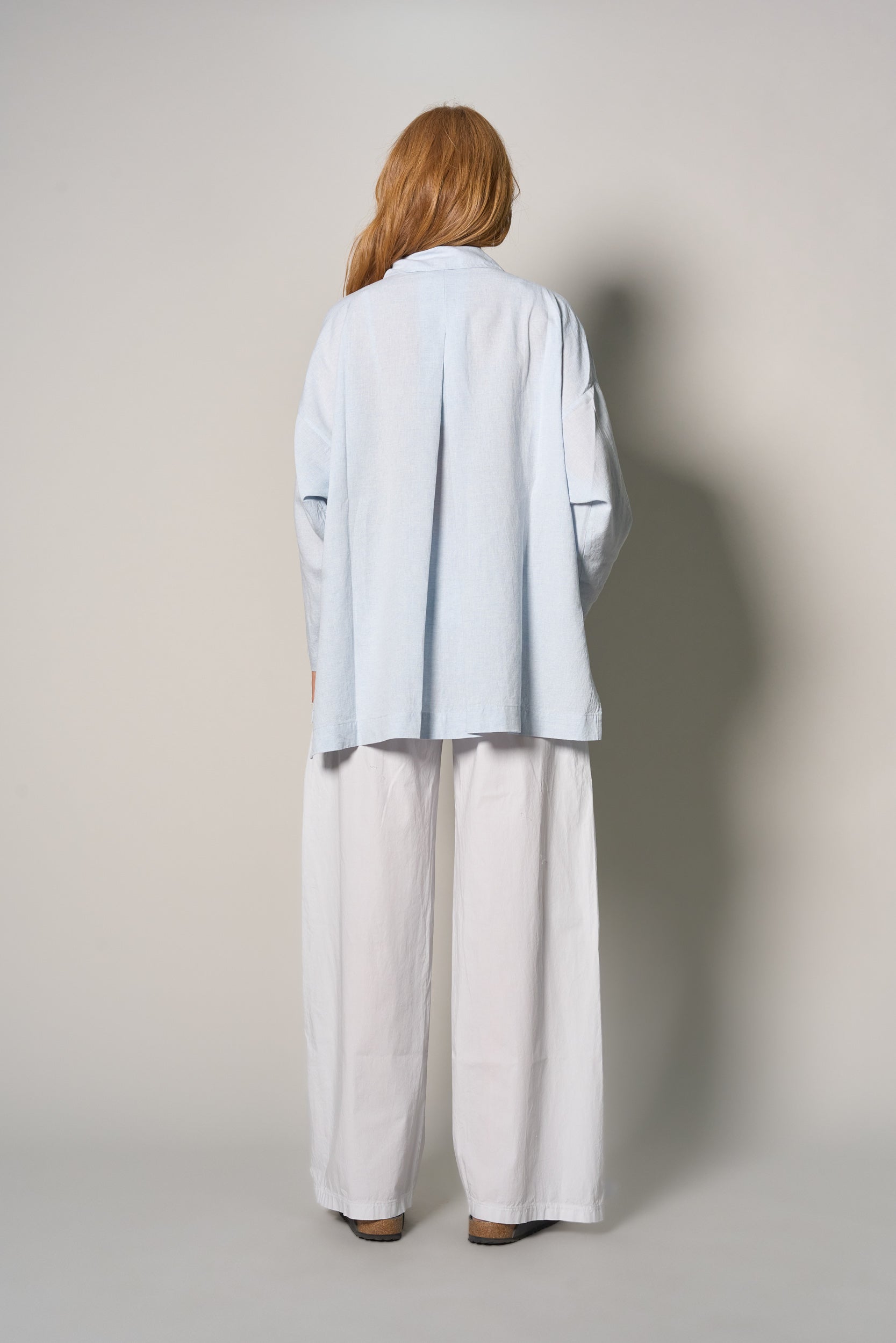 A model viewed from behind wearing a light blue oversized button-down shirt and white wide-leg pants, standing against a neutral light gray studio background.