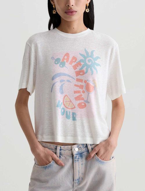 A woman wearing a white cropped t-shirt with a colorful retro graphic that reads 'APERITIVO HOUR' with a cocktail glass, sun, and citrus slice, paired with light-wash jeans against a plain light gray background.