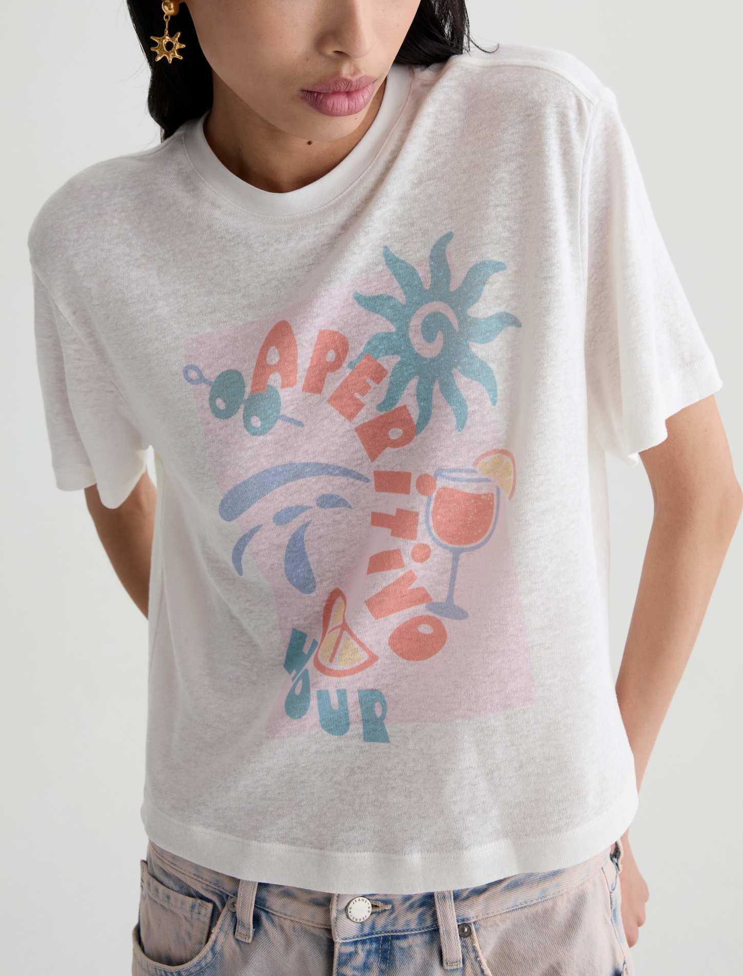 A close-up of a model wearing a white heathered graphic t-shirt with a colorful retro design featuring the words 'APRÈS L'EAU' and summer-themed illustrations, paired with light-wash jeans.