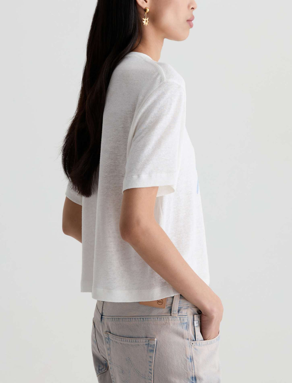 A woman shown from the side wearing a white short-sleeve cropped t-shirt and light-wash jeans against a plain light gray background.